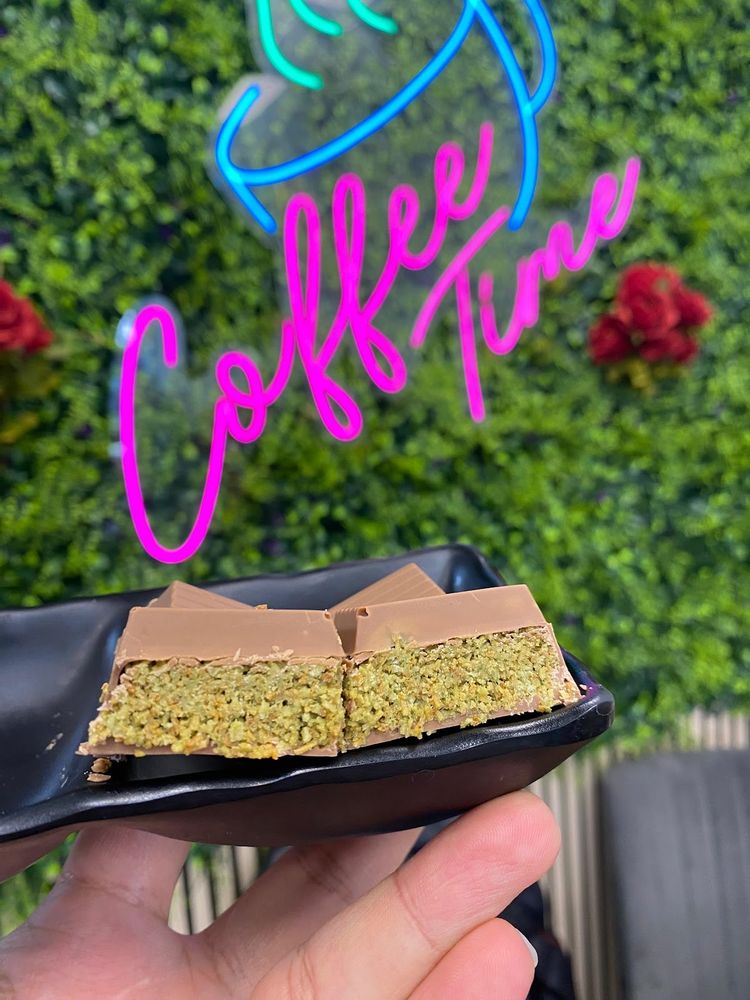 A hand holding a plate of two slices of chocolate-topped pistachio traybake